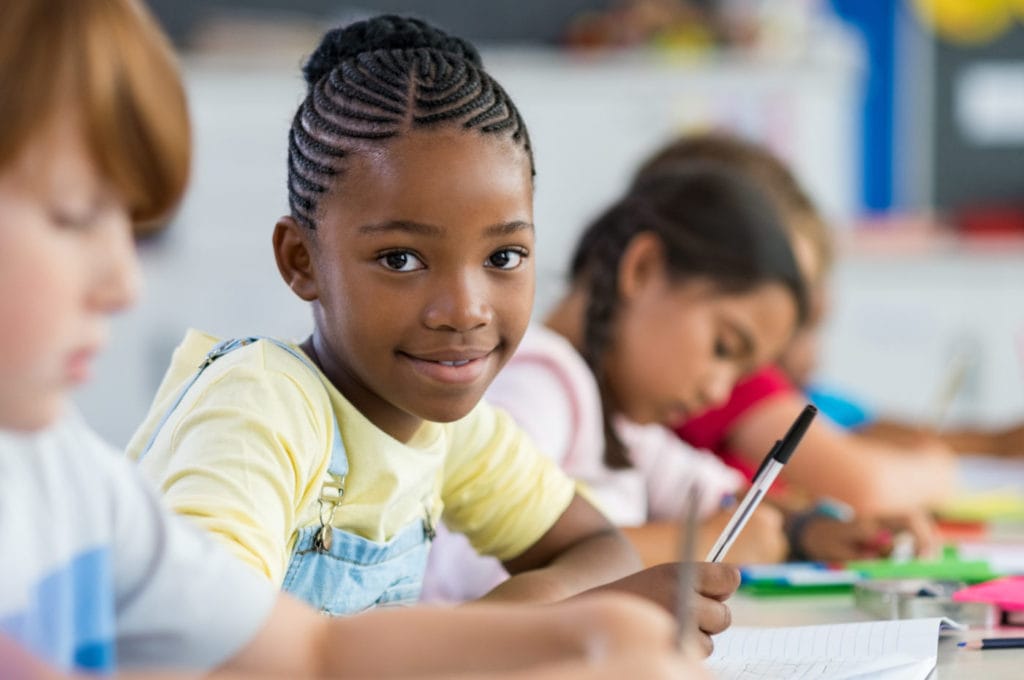 Smiling african girl sitting at desk in class room and looking at camera. Portrait of young black schoolgirl studying with classmates in background. Happy smiling pupil writing on notebook.